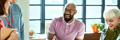 Mid adult African man in glasses with coffee, smiling and talking to colleague, senior woman using laptop and making notes